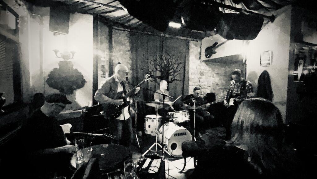 Black and white image of a live band rehearsal session with musicians performing together in a rehearsal room for Music Scene Magazine’s “So You Wanna Build a Band?” series.