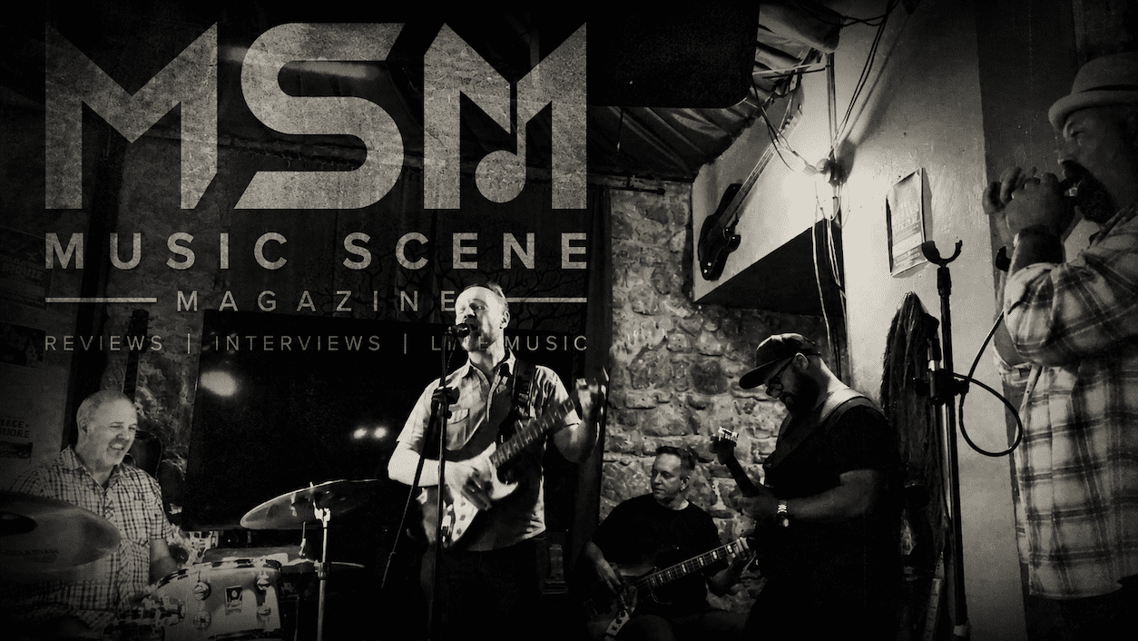 Black and white live blues performance image featuring musicians performing on stage with Music Scene Magazine branding overlay.