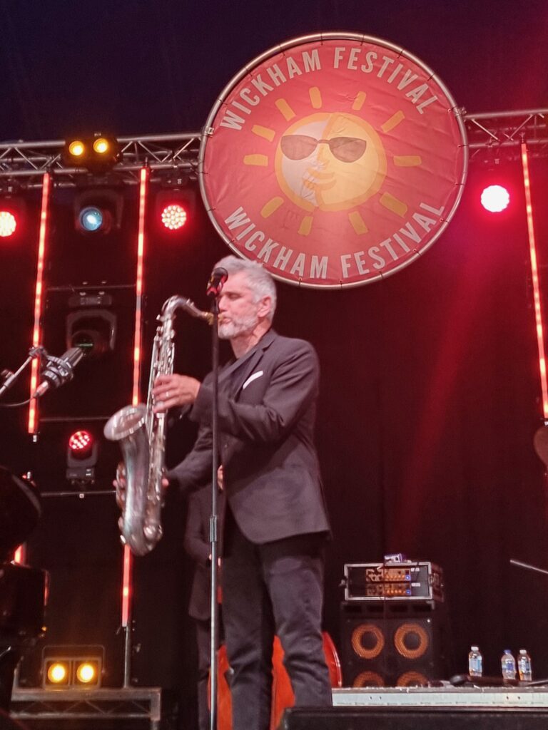 A saxophonist performing live on stage beneath the Wickham Festival logo. Image supplied courtesy of Wickham Festival.